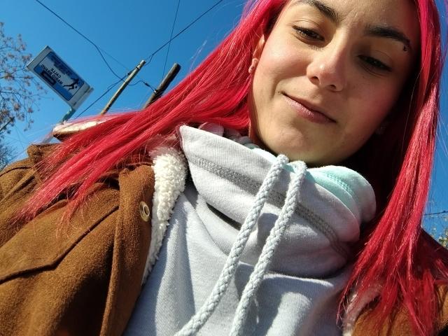 AmyStreetwise69 with red hair, brown coat, gray hoodie, and white drawstring, smiling slightly, blue sky background, brown eyes, light skin, and small mole above her right lip.