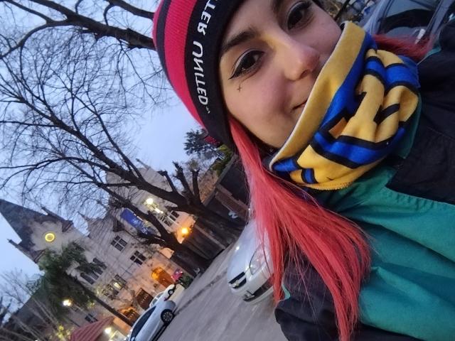 AmyStreetwise69 with bright pink hair, green jacket, red beanie, yellow and blue mask, holding it over her mouth, standing outside in a snowy urban setting.