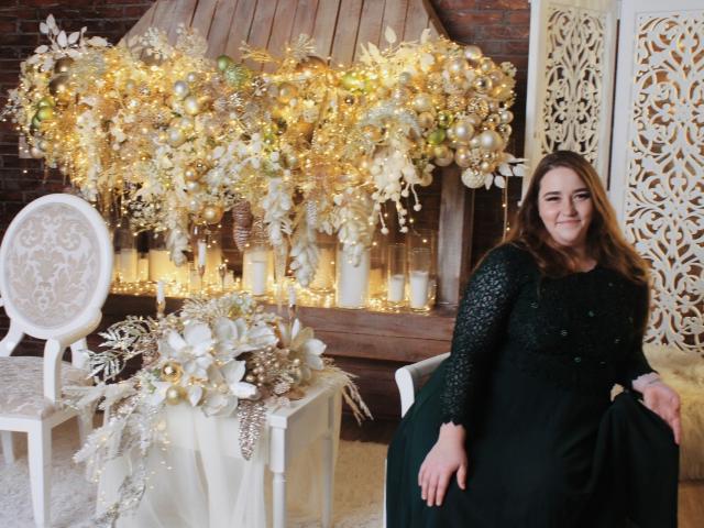 DaniellaGraceZZ has tits in a dark green dress with a lace top, standing next to a white chair with a floral-patterned seat. She's smiling, with a gold and white decor.
