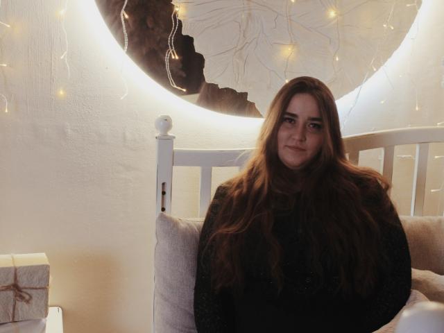 DaniellaGraceZZ with long brown hair, black top, sitting on white bed, smiling, soft light, fairy lights, pillow, gift wrapped on left, white headboard, cozy, warm atmosphere.