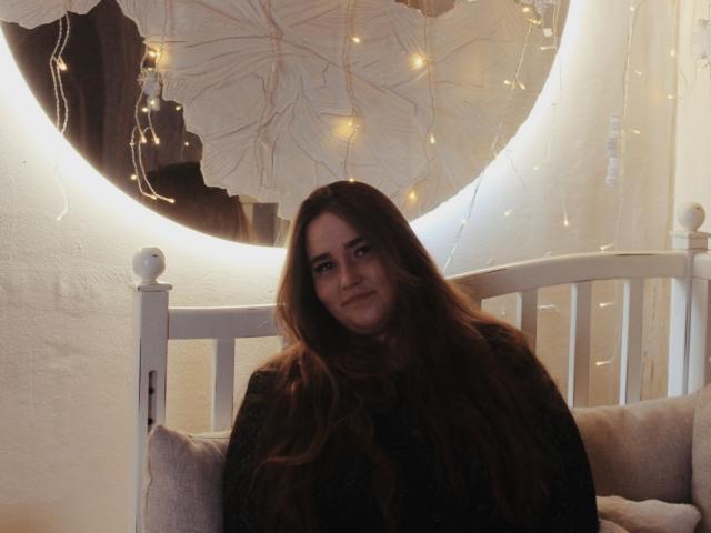 DaniellaGraceZZ con cabello castaño largo, suéter oscuro, sentada en una cama blanca, con una sonrisa suave, luces de hadas en el fondo, un adorno circular blanco en la pared, un ambiente acogedor y cálido..