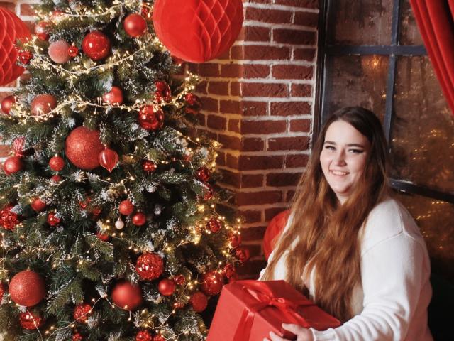 DaniellaGraceZZ with long brown hair, white sweater, holding red gift, smiling next to decorated Christmas tree with red ornaments and lights.