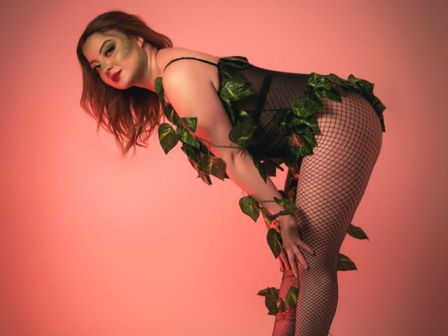RedSoull has green leaves wrapped around her black mesh lingerie and fishnet legs, bending forward with red lipstick and green eye makeup, showing her ass and looking at the camera.
