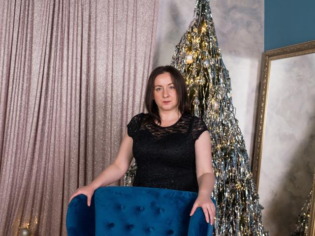 AbbyTaylorr with tits in a black lace dress, sitting on a blue velvet chair, hands on arms, next to a glittery Christmas tree, smiling, brown hair, blue eyes.
