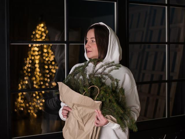 AbbyTaylorr with brown hair, white hoodie, holding brown bag with green pine branches, standing in front of black-framed window with blurred golden Christmas lights in background.