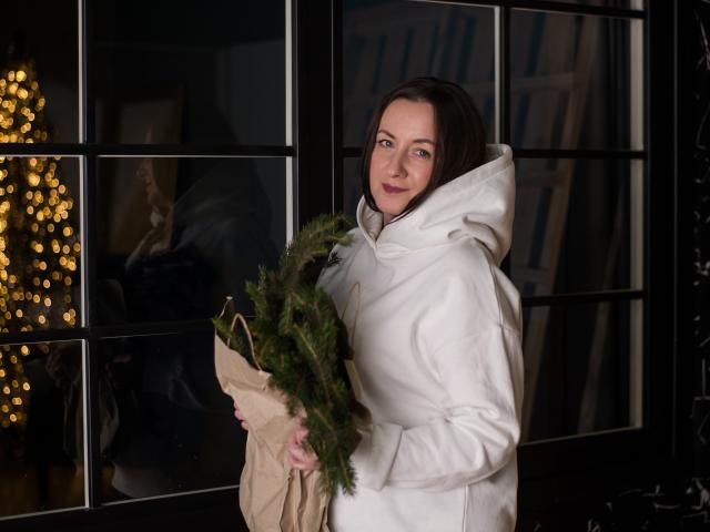 AbbyTaylorr with short brown hair, white hoodie, holding brown bag with greenery, standing by black-framed glass door, looking at camera with slight smile, blue eyes, soft lighting.