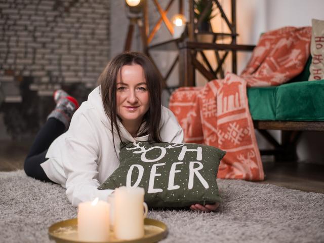 AbbyTaylorr, laying on a grey rug, smiling with brown hair, white hoodie, holding a green pillow, candles in front, Christmas decor, green velvet chair, brick wall background.