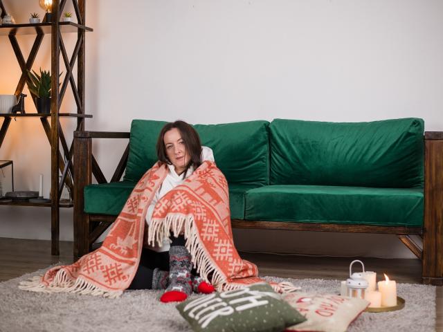 AbbyTaylorr with medium brown hair, sitting on green couch, wrapped in orange patterned blanket, smiling, red pom-poms, white shirt, wooden floor, candles, gray rug, green pillow.