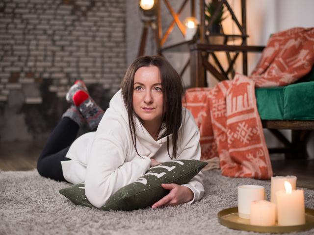 AbbyTaylorr laying on a gray rug, white sweater, green pillow, black socks with red and gray patterns, candles in the foreground, cozy indoor setting, brown hair, cute smile.