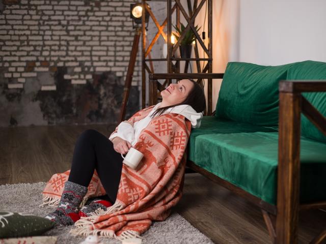 AbbyTaylorr, with tits covered, sits on floor, legs crossed, wearing orange patterned blanket, green socks, white shirt, black pants, and green sofa, smiling, holding white mug.