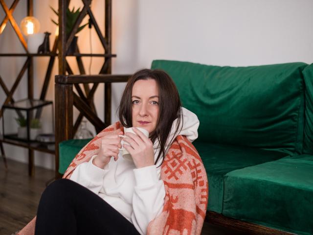 AbbyTaylorr with medium tits, brown hair, white hoodie, orange patterned blanket, green velvet couch, holding white mug, black pants, wooden shelves, warm light, wooden floor.