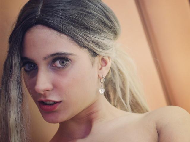 DelaMoons mit blonden Haaren, blauen Augen und herzförmigen Ohrringen, die in die Kamera schauen mit leicht geöffneten Lippen und dabei ihre nackten Schultern zeigen. Ihr Gesicht hat einen dezent rosafarbenen Schimmer..