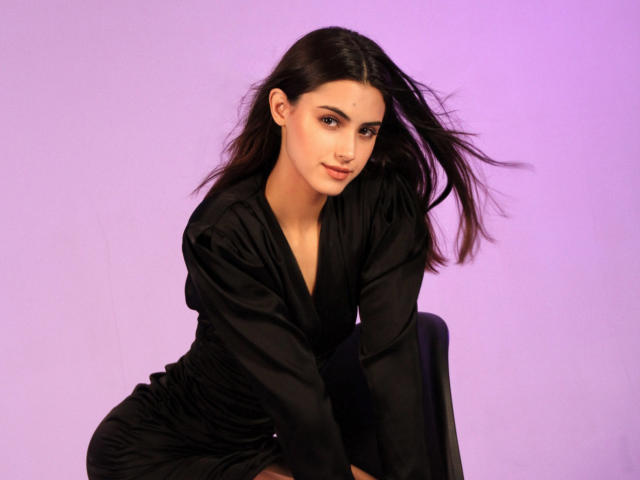 JasmineDay a les cheveux foncés, porte une robe noire et a un joli visage. Elle est assise avec un léger sourire, regardant l'appareil photo, et ses seins sont subtilement visibles sous la robe..