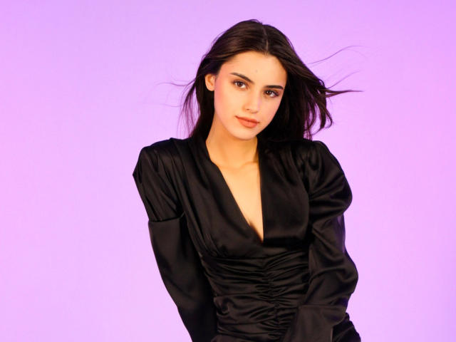 JasmineDay with long dark hair, wearing a black satin dress with a deep V-neck, sitting against a purple background, looking slightly to the side with a gentle smile.