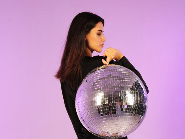 JasmineDay holding a shiny silver disco ball, wearing a black long-sleeve top, with medium-length dark brown hair, purple lighting, and a focused expression.