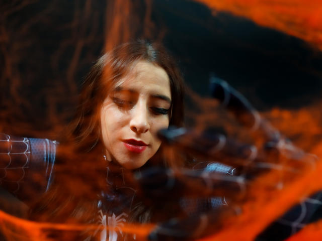 Kaiylin mit rotem Lippenstift und braunen Haaren, im Hintergrund verschwommene orange und schwarze Wirbel, trägt ein glänzendes, dunkles Kleidungsstück und blickt mit einem leichten Lächeln nach unten..