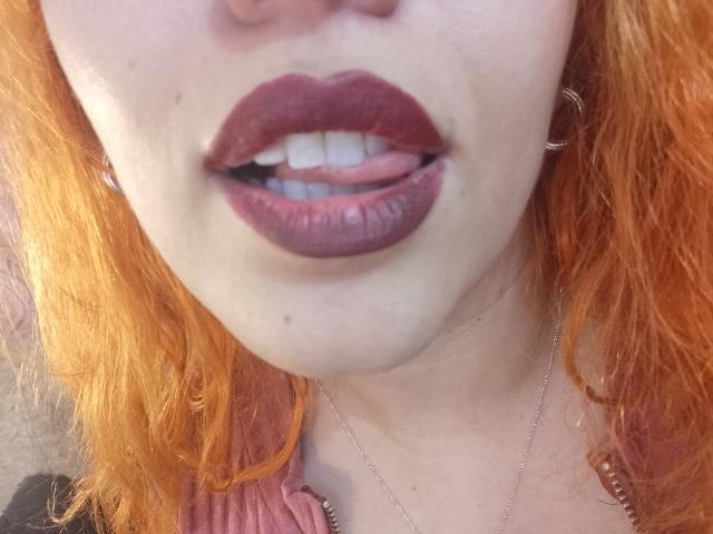 Het gezicht van Joanallen, met rood-oranje krullend haar, volle roze-bruine lippen met donker paarse lippenstift, lichtjes open met tanden zichtbaar, draagt een roze top en een zilveren ketting..