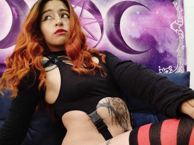 YulikoOfficial with red hair, black cutout top, black arm sleeves, tattoo on right side, striped red and black socks, looking up with surprised expression, purple moon tapestry background.