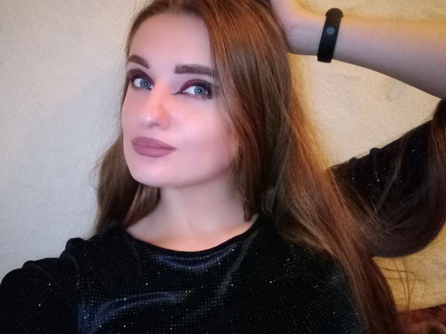 KariMillie, con cabello castaño largo, ojos azules y lápiz labial rosa, vestida con una blusa negra brillante, con la mano en el cabello, una pulsera negra, mirando ligeramente hacia un lado, con una sonrisa suave..