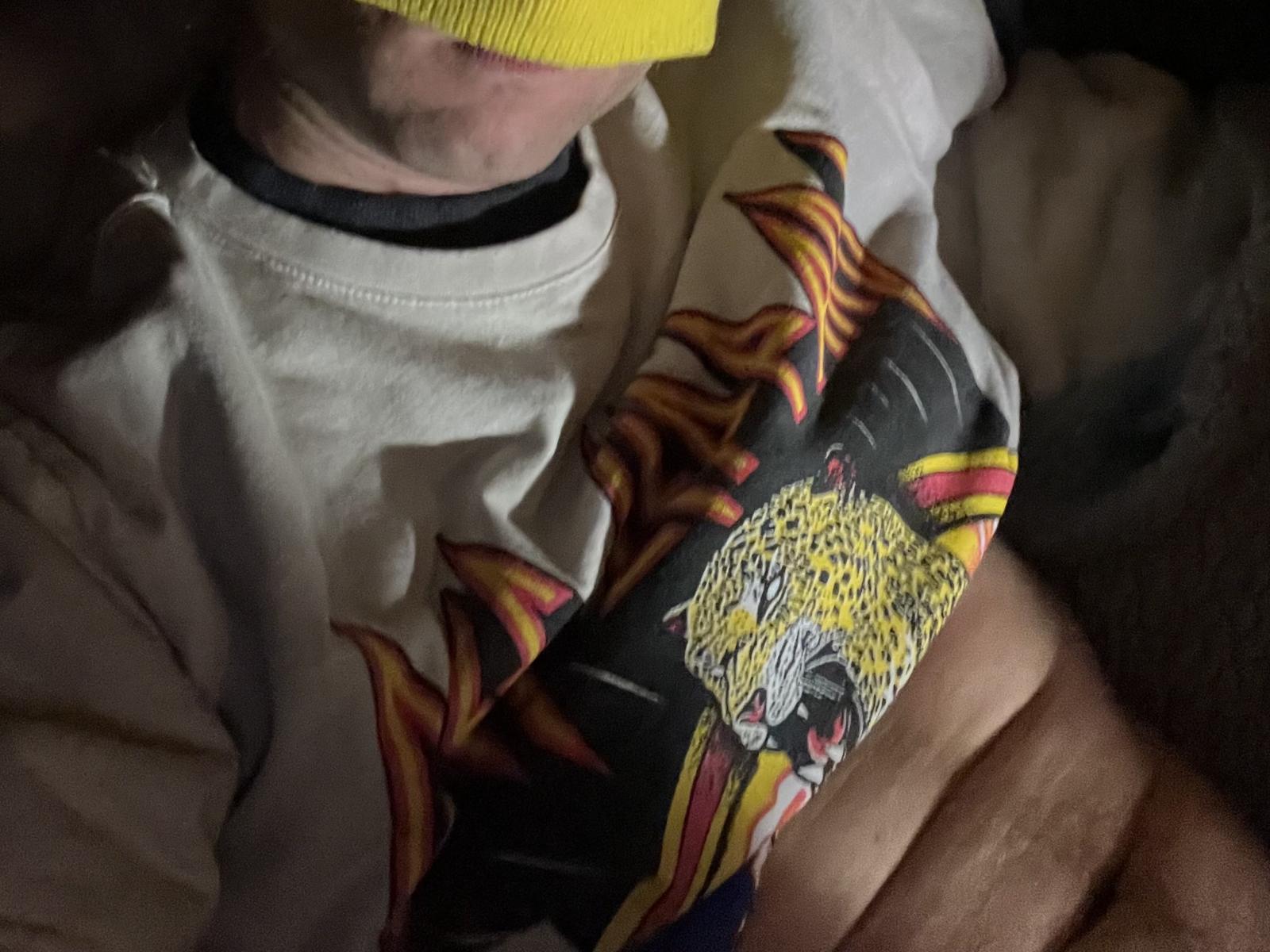 Metusela con una máscara amarilla, una camisa blanca con un estampado colorido de un leopardo, y los brazos cruzados. La camisa tiene llamas a los lados. Con poca luz, enfocada en la parte superior del cuerpo..