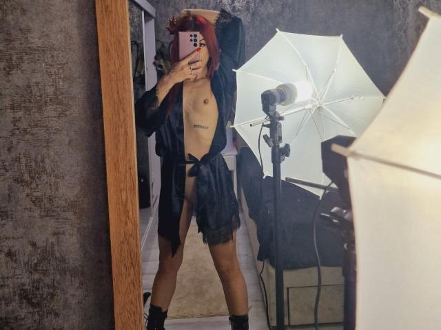RinaQueenie met rood haar, naakt behalve een zwart shirt dat in de taille is vastgebonden, zwarte kanten shorts en laarzen, staat voor een spiegel en houdt een roze telefoon vast..