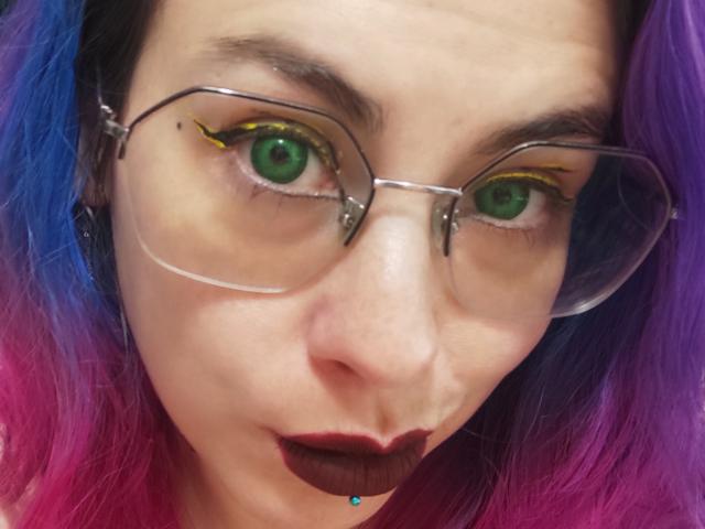 NatashaLau with green eyes, wearing clear glasses, has purple and blue hair, dark lipstick, and a small piercing below her chin. Her makeup includes yellow eyeliner.