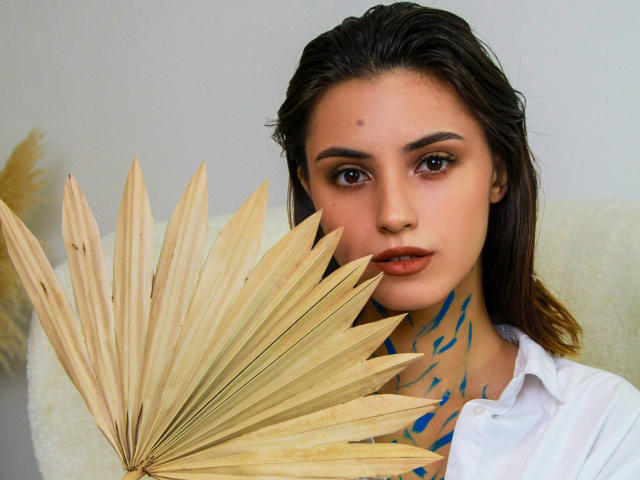 JasmineDay, avec une peau claire, des yeux marron foncé et des tatouages bleus sur le cou, tenant un éventail avec des feuilles beige, portant une chemise blanche, regardant directement l'appareil photo..