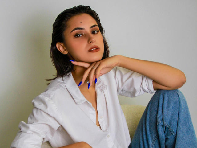 JasmineDay with dark hair, wearing a white button-up shirt and blue jeans, has blue nails and rests her chin on her hand, looking sexy with a slight smile.
