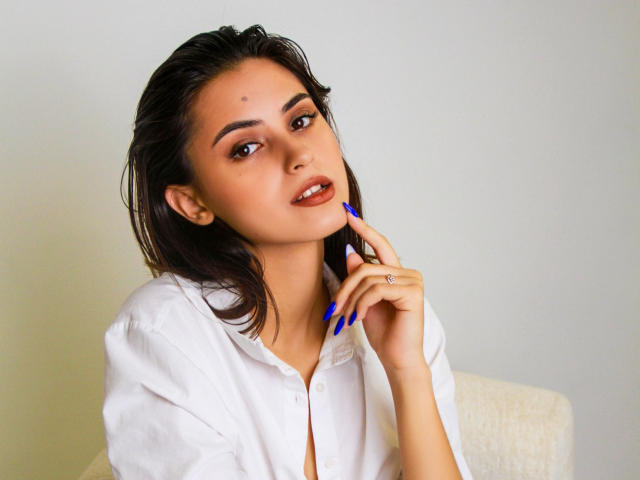 JasmineDay, con la pelle marrone media, i capelli scuri e umidi e le unghie blu, indossa una camicia bianca, tocca il mento con un anello al dito e guarda la fotocamera..
