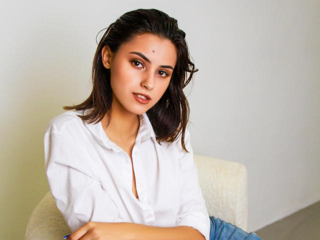 JasmineDay has a cute face, medium tits, dark brown hair, and is wearing a white button-up shirt with blue jeans, sitting with arms crossed, looking at the camera.