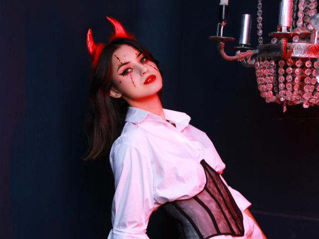 JasmineDay with red devil horns, black makeup, red lipstick, white shirt, black corset, and dark hair, standing near a crystal chandelier, looking up with a seductive smile.