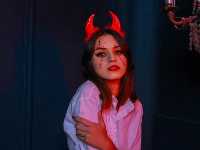 JasmineDay con corna da diavolo rosse, sangue sul viso, rossetto rosso, camicia bianca, con un braccio alzato, sfondo blu scuro, illuminazione rossa e blu, candelabro a destra..