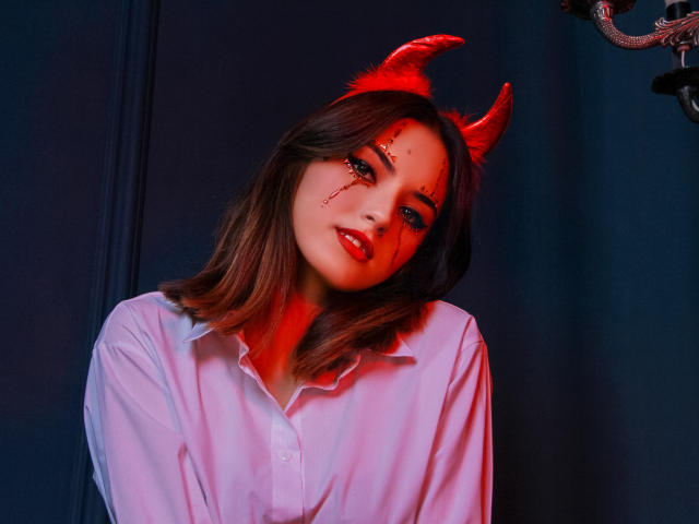 JasmineDay with red devil horns, bloody face makeup, and red lipstick, wearing a white button-up shirt, has medium tits and wavy brown hair, looking seductive in red light.