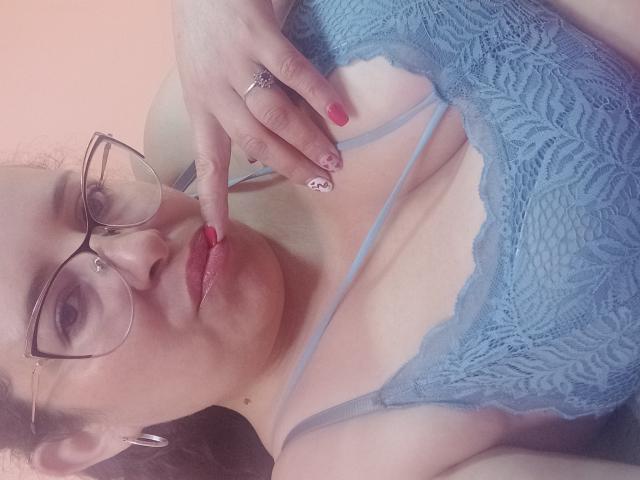 CandHoney con gli occhiali, reggiseno di pizzo blu, dito vicino alla bocca, smalto rosso, anello, e un viso carino, che mostra il seno e un'espressione sexy..