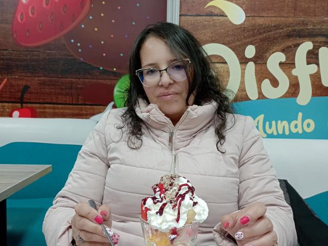 CandHoney with curly dark hair, glasses, pink puffy jacket, holding spoon and fork, pink nail polish, dessert with whipped cream, strawberries, and chocolate in a glass.
