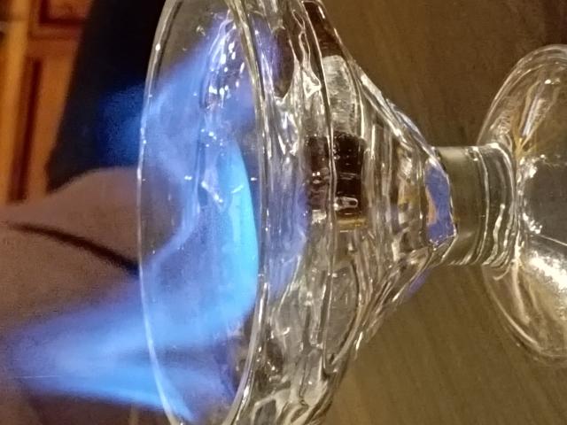 Bougeoir en verre transparent avec une flamme bleue à l'intérieur, mettant l'accent sur la lueur de la flamme et le design texturé et facetté du bougeoir. La main de CandleHoney est partiellement visible sur la gauche..