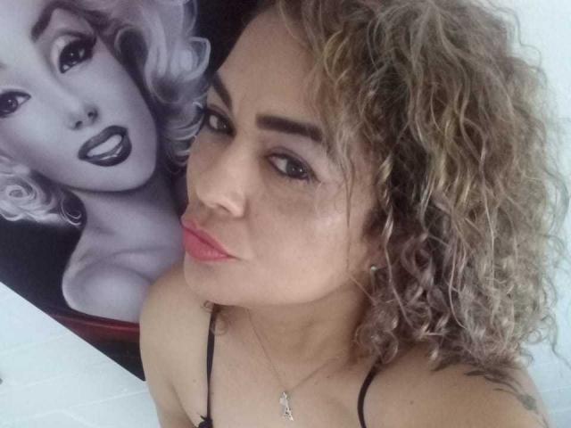 SolmaryHot with curly blonde hair, pink lipstick, and black spaghetti strap top, pouting lips, black eyeliner, and necklace, standing next to a Marilyn Monroe portrait.