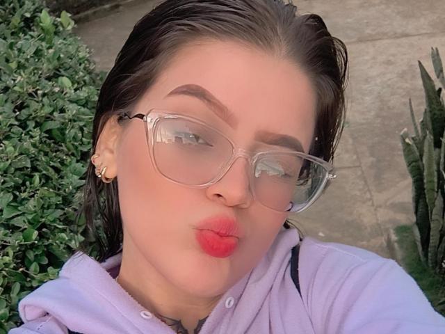 KimSummer with wet brown hair, clear glasses, red lipstick, and a purple hoodie, pouting her plump, shiny-assed lips. She's wearing gold earrings and has a cute face.