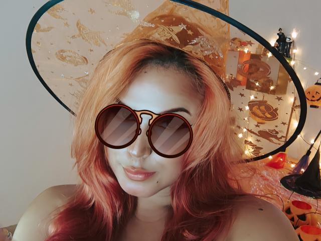AhriValvet, con cabello rosado-rojizo, usando un sombrero de bruja y gafas de sol redondas, mostrando sus pechos, sonriendo a la cámara. Tiene el nose perforado. Se ve luz de velas en el fondo..