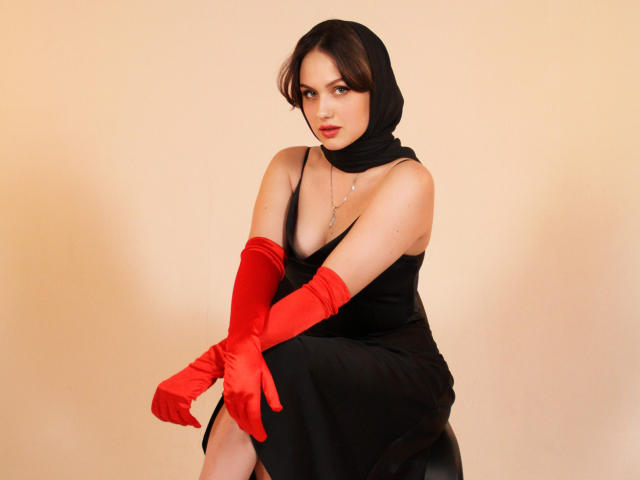 Lisa Francis trägt ein schwarzes Kleid mit roten Handschuhen, einem schwarzen Kopftuch und einer Halskette. Sie sitzt mit einem verführerischen Ausdruck, wobei ihre Brüste und ein Schlitz im Bein zu sehen sind, und wirkt sehr attraktiv..