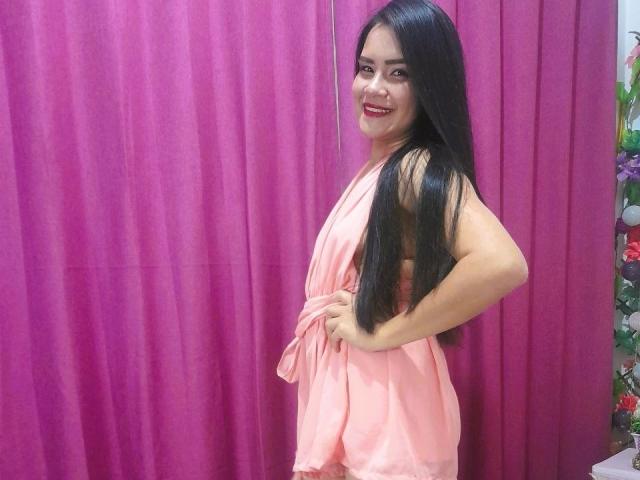 ConniRiccy, con cabello negro largo, sonriendo, vestida con un vestido de satén rosa, con las manos en las caderas, de pie de lado, mostrando su trasero, fondo de cortina rosa, lápiz labial rojo, luciendo linda..