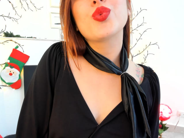 LaiaReyd con labbra rosse carnose, vestito nero, scollatura a V profonda, sciarpa nera legata al collo, tatuaggi visibili sulla spalla, capelli castani, sfondo bianco con decorazioni natalizie..