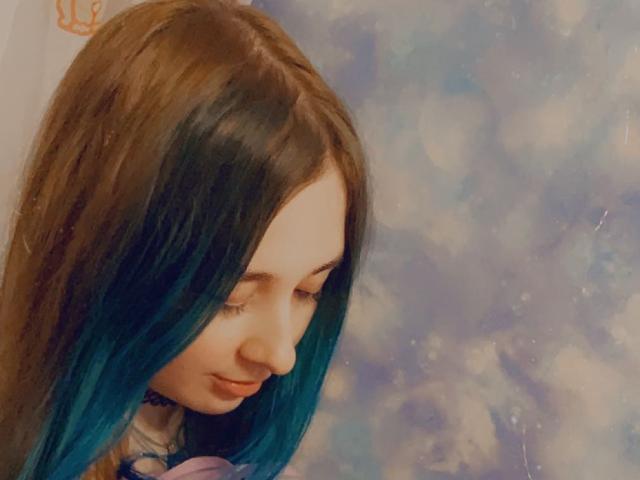 HentaiShing69 hat langes, braunes Haar mit blauen Spitzen, ein blasses Gesicht und eine kleine Nase und blickt mit einem leichten Lächeln nach unten. Ihr Hintern ist nicht sichtbar..