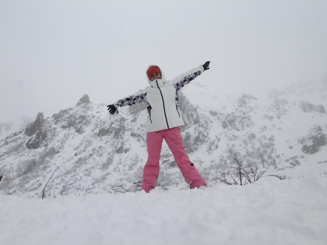 DelaMoons è in piedi nella neve, con le braccia aperte, indossa una giacca bianca, maniche con motivi floreali, pantaloni rosa e un cappello rosso. È circondata da neve bianca e cime rocciose..