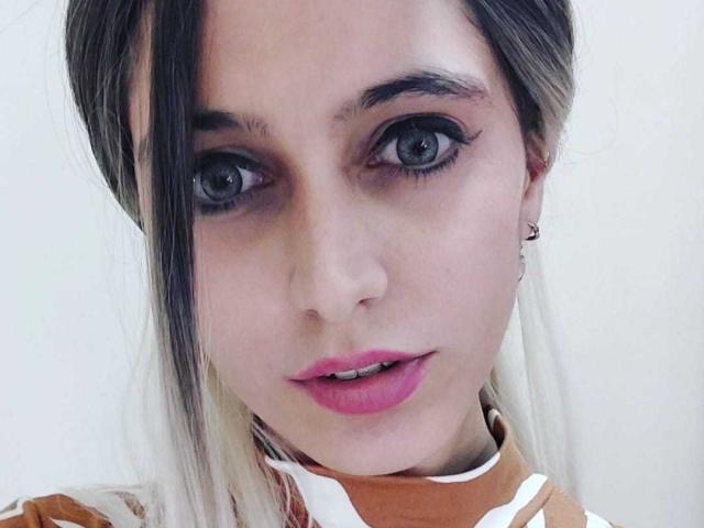 DelaMoons tiene grandes ojos azules, labios rosados y una boca ligeramente abierta. Tiene cabello rubio y negro, y lleva una blusa marrón y blanca, con un pequeño arete..