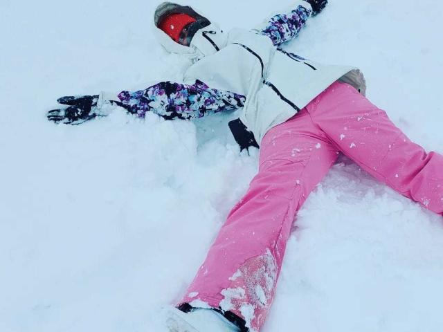 DelaMoon liegt bäuchlings im Schnee, die Arme ausgestreckt, und trägt eine weiße Jacke, violett-schwarze Ärmel, pinke Schneehosen und rote Skibrillen..