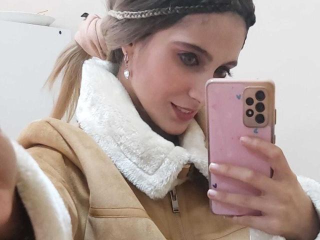 DelaMoons con i capelli castani raccolti in una treccia, indossa un cappotto color sabbia con pelliccia bianca, tiene in mano un telefono rosa, sta scattando un selfie, sorride, mostrando un viso carino e unghie rosa..