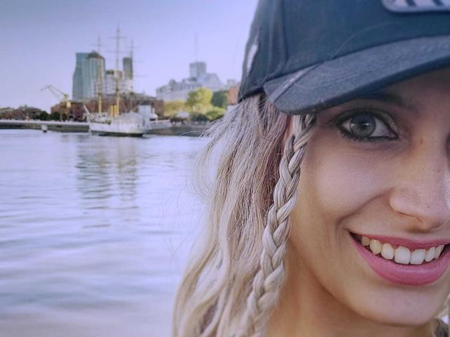 DelaMoons, com cabelo loiro trançado, olhos azuis e batom rosa, usando um boné preto, sorrindo para a câmera, mostrando dentes brancos e um pequeno piercing no nariz. Água ao fundo..