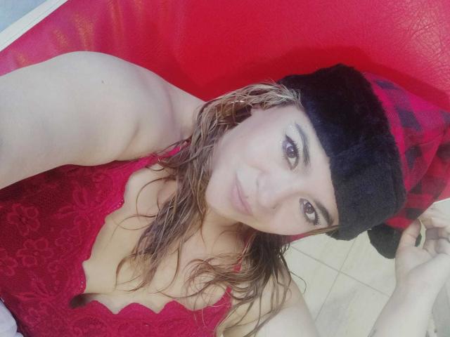NaugthyCurvy, com cabelos loiros molhados, vestindo uma blusa vermelha de renda e um gorro preto/cinza, tem olhos castanhos e um leve sorriso, exibindo seus seios em uma selfie..