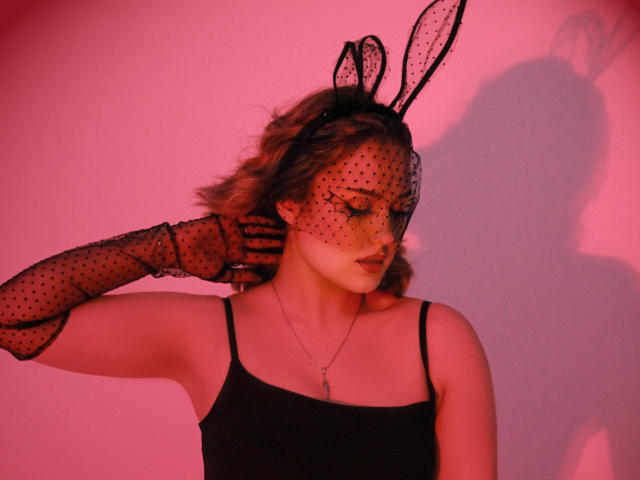 Lisa Francis portant un masque de lapin noir à pois avec des oreilles, une robe noire à bretelles, et des gants assortis à pois, une lumière rouge projetant une ombre, des cheveux bruns ondulés, un collier avec une croix..